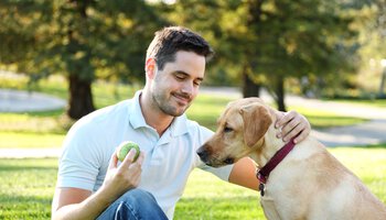 Ein Mann mit einem Tennisball in der Hand sitzt auf dem Boden/Rasen und streichelt einen Hund | © Gene Chutka - Getty Images